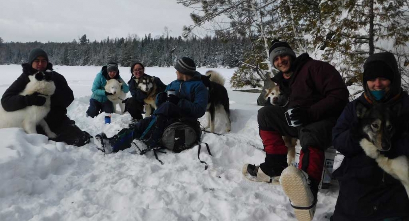 A group of people wearing heavy snow gear smile at the camera while they cuddle with sled dogs in a snowy landscape